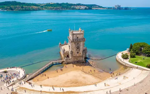 Wieża Belém, symbol kultury Portugalii, dumnie stoi nad wodą. Turyści spacerują po plaży i nabrzeżu.