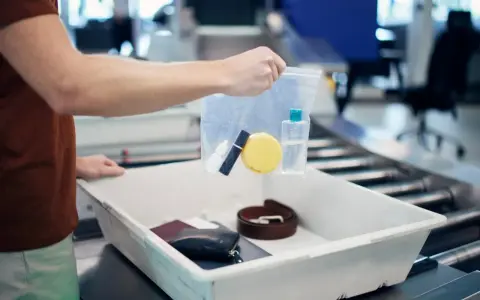 Ręka trzyma przezroczystą torebkę z kosmetykami nad tacą z rzeczami do kontroli bezpieczeństwa na lotnisku.