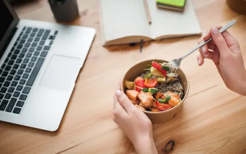 Ręce trzymające miskę z łososiem i warzywami, obok laptop i notatnik. Idealny posiłek na dieta okienkowa.
