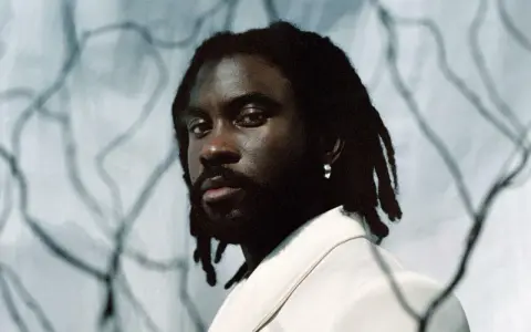Retrato de un rapero español con rastas y un pendiente plateado, vistiendo una chaqueta clara.