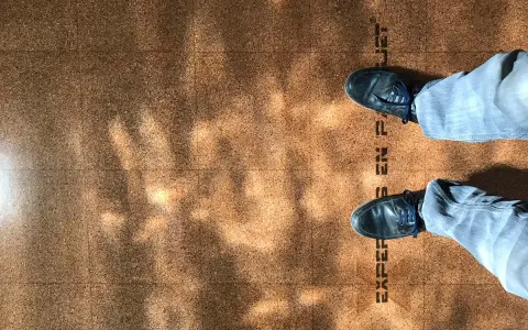 Pintura para corcho con un diseño de baldosas y sombras de luz. Se ven unos pies con zapatos oscuros y pantalones vaqueros.