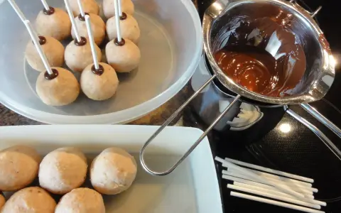 Biskuit-Cake-Pops warten auf ihre Schokoladenhülle. Geschmolzene Schokolade und Stäbchen liegen bereit.
