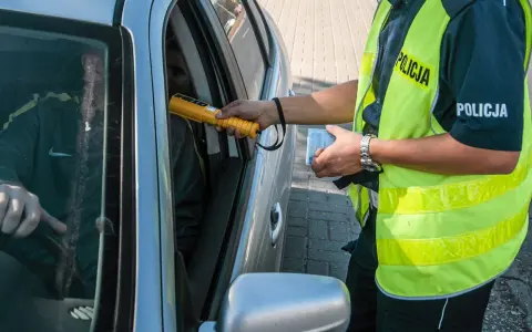 Policjant bada kierowcę alkomatem, sprawdzając, czy nie ma **alkoholu w organizmie**. Kierowca siedzi za kierownicą, a funkcjonariusz trzyma urządzenie przy jego twarzy.