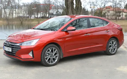 Czerwony Hyundai Elantra 2023 zaparkowany nad jeziorem, z drzewami i budynkami w tle.