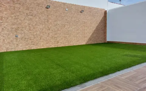 Un patio con césped artificial verde vibrante, paredes de piedra y madera. Ideal para aprender como colocar césped artificial.