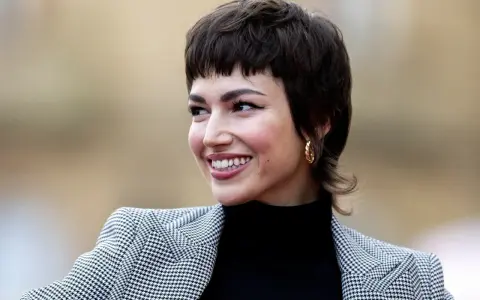 Uśmiechnięta kobieta z charakterystyczną fryzurą bixi, w czarnym golfie i wzorzystej marynarce.