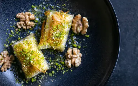 Zwei Stück Baklava, bestreut mit gehackten Pistazien und Nüssen, auf einem dunklen Teller. Ein wahrhaftiges Baklava Rezept Original.