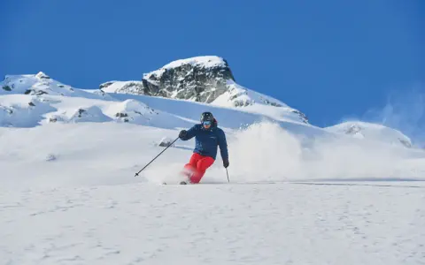 Skifahrer in roter Hose und blauer Jacke wirbelt Schnee auf. Die Columbia Omni-Heat Erfahrung: warm und trocken im Pulverschnee.
