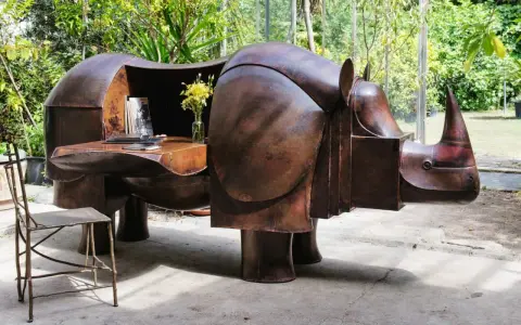 Étonnante sculpture lalanne en forme de rhinocéros, bureau fonctionnel en cuivre patiné, avec un vase de fleurs jaunes et une chaise en métal.