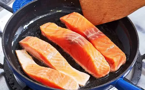 Vier Lachsfilets braten in einer Pfanne. Ein Holzspatel liegt bereit, um zu zeigen, wie bereite ich Lachs zu.