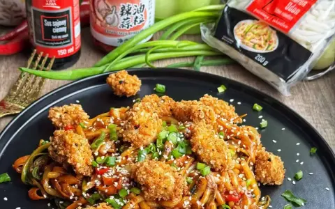 Pyszny makaron udon z kurczakiem w panierce i kolorowymi warzywami, posypany sezamem i szczypiorkiem.