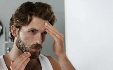 Mann mit unreiner Haut reinigt sein Gesicht mit Schaum. Er blickt in den Spiegel, während er seine Stirn einreibt.