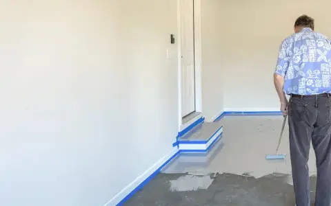 Mann beschichtet den garagenboden mit einem Roller. Der Boden ist teilweise mit einer grauen Beschichtung bedeckt, die Ränder sind mit blauer Klebefolie abgeklebt.