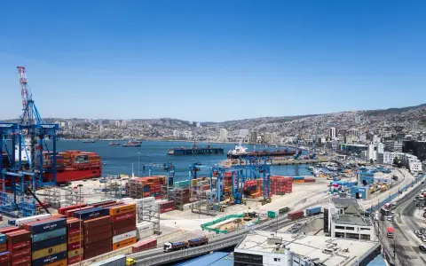 Un vasto porto con gru, container colorati e navi cargo, con una città collinare sullo sfondo. Sembra il porto più grande del mondo.