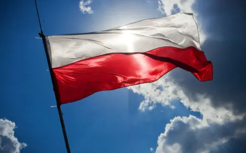 Biało-czerwona flaga Polski powiewa na tle błękitnego nieba z chmurami. Słońce przebija się przez materiał, podkreślając jego fakturę. Wymiary flagi polski są standardowe, proporcje zachowane.