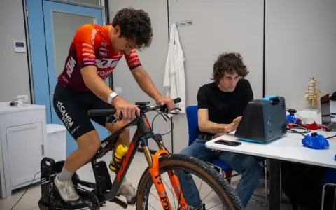 Ciclista su rulli monitorato per l'acido lattico, con un tecnico che osserva i dati su un dispositivo.