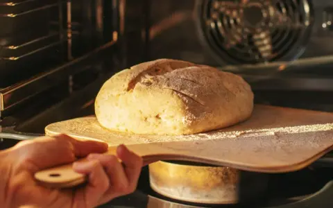 Ręka wsuwa drewnianą łopatą bochenek chleba do rozgrzanego piekarnika. To kulminacyjny moment wyrabiania ciasta na chleb.