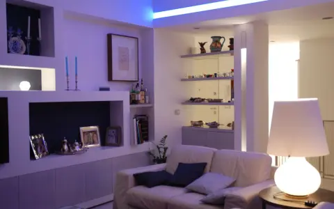 Soggiorno moderno con illuminazione soffusa, ideale per come illuminare un soggiorno con poca luce. Divano bianco, mensole con oggetti e lampada da tavolo.