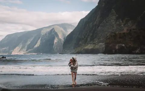 Kobieta z dzieckiem na rękach na plaży, w tle majestatyczne góry i ocean. Niezapomniane madera widoki.