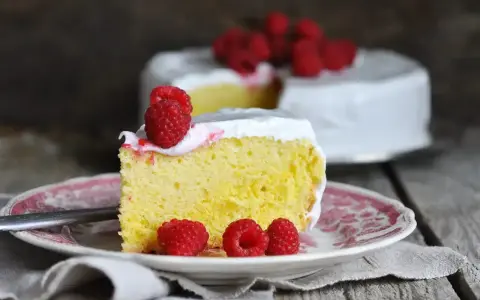 Wilgotne ciasto tres leches z malinami na talerzyku, w tle cały tort.
