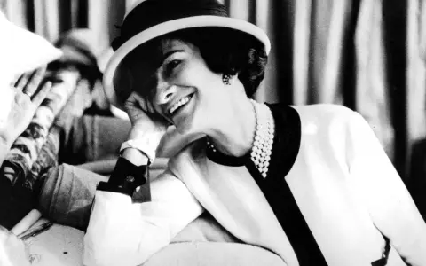 Coco Chanel, sonriendo con un sombrero y perlas, evoca la elegancia de donde es Chanel.