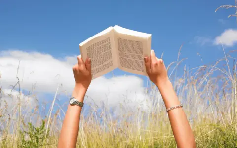 Mani che sorreggono un libro aperto contro il cielo azzurro, tra fili d'erba alta. Un momento di pace e apprendimento, magari con libri sulla salute mentale.