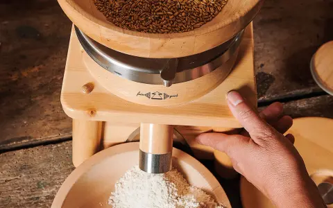 Holzgetreidemühle mit Getreide oben und frisch gemahlenem Mehl unten, Hand bedient die Mühle.