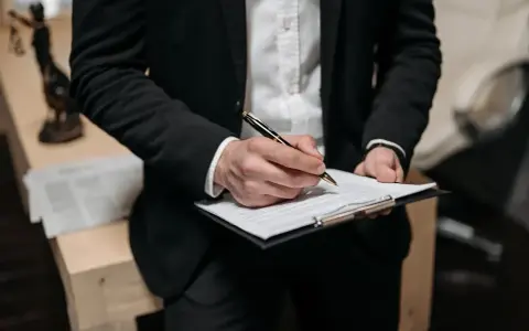Prawnik w czarnym garniturze podpisuje pozew o zapłatę na dokumencie. W tle widać figurkę Temidy.