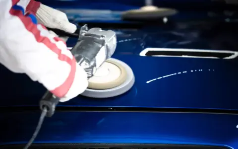 A gloved hand uses a buffer to polish a shiny blue car hood, demonstrating how to polish a car with a buffer for a flawless finish.