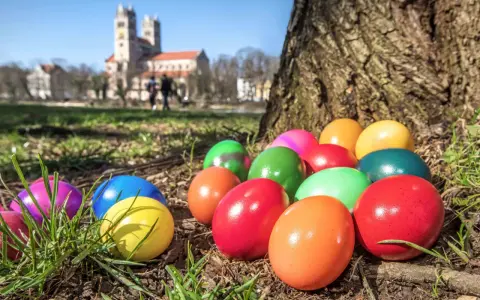 Bunte Ostereier liegen im Gras vor einem Baum. Im Hintergrund ist eine Kirche zu sehen. Die Suche nach den Eiern ist ein schöner Teil der Ostern Bedeutung.