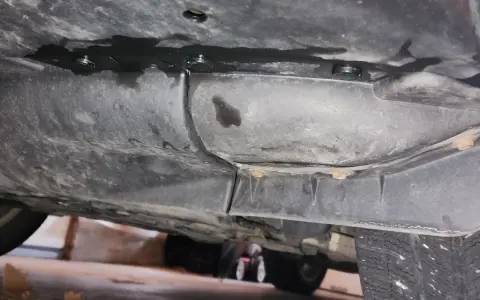Underneath a car, dark fluid is leaking from a seam. A small puddle of blue fluid is visible, suggesting a coolant leak.