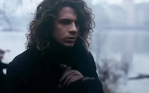Mężczyzna z kręconymi włosami w czarnym płaszczu, trzymający się za rękę, jakby chciał powiedzieć "never tear us apart".