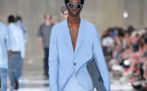 Modello con outfit uomo elegante senza giacca, completo azzurro, occhiali da sole blu e borsa a righe.