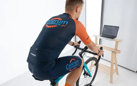 Mężczyzna w stroju kolarskim pedałuje na rowerze stacjonarnym, śledząc trasę na ekranie laptopa z aplikacją Zwift.