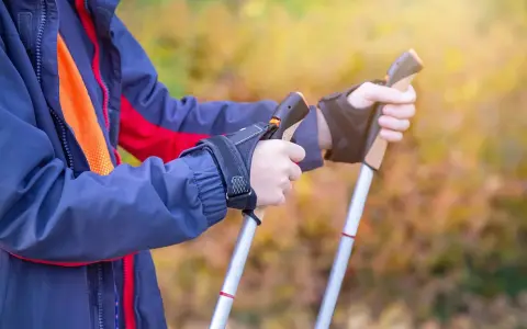 Osoba w kurtce ćwiczy, jak chodzić z kijkami trekkingowymi. Dłonie pewnie trzymają uchwyty, gotowe do marszu.