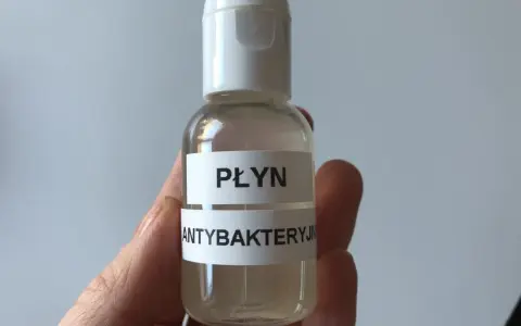 Dłoń trzyma małą, przezroczystą butelkę z białym zamknięciem, zawierającą płyn.