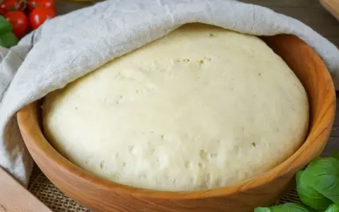 Frischer Pizzateig ruht in einer Holzschüssel, bedeckt mit einem Tuch. Tomaten und Basilikum im Hintergrund deuten auf die bevorstehende Zubereitung hin.
