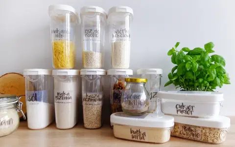 Pojemniki na żywność z etykietami pokazują, jak przechowywać pokrywki i produkty sypkie. Widać makaron, ryż, sól, mąkę, kaszę, cukier puder i płatki owsiane.