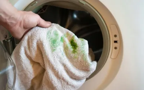 Hand hält Handtuch mit grünen, eingewaschenen Flecken vor einer Waschmaschine.