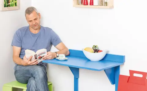 Mann liest Buch an selbstgebautem Klapptisch. Obstschale und Kaffeetasse stehen bereit.