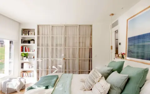 Estantería blanca aprovecha hueco entre cama y pared, con libros y plantas. Puertas de cristal con cortinas separan el espacio.