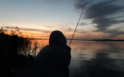 Zachód słońca nad jeziorem, wędkarz czeka na branie. Zastanawia się, jaka będzie karta wędkarska cena.