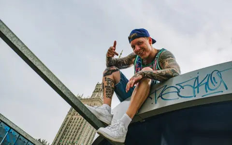 Uśmiechnięty raper z tatuażami, w czapce i kolorowej koszulce, siedzi na dachu, promując swój mixtape "sos ciuchy i borciuchy".