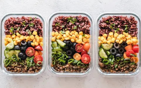Drei Glasbehälter mit einem gesunden Essensplan für Fitness: Linsen, Gurken, Oliven, Tomaten, Mais und brauner Reis.