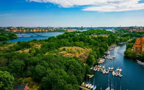 En vacker promenad i Stockholm med grönskande träd, en bro över vattnet och många segelbåtar i hamnen.