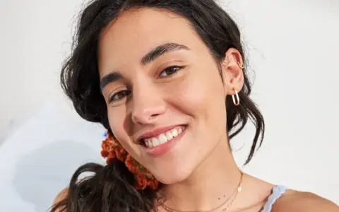 Primo piano di una giovane donna con un sorriso radioso, la sua fronte lucida riflette la luce. Indossa orecchini a cerchio dorati e una collana sottile.