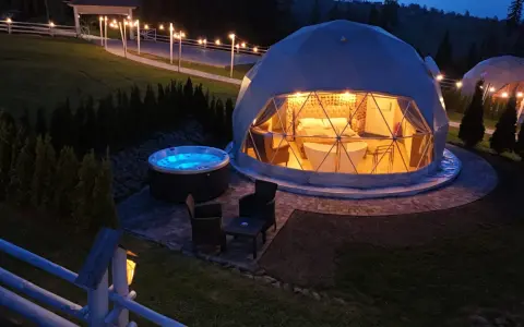 Nocny widok na luksusowy camping w Tatrach. Dwa kopułowe namioty, jeden z wanną i łóżkiem, drugi z oświetleniem. Obok jacuzzi i meble ogrodowe.