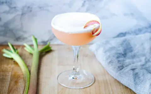 En rosa rabarber sour-drink med skum och en rabarberdekoration. Två stjälkar rabarber ligger bredvid.