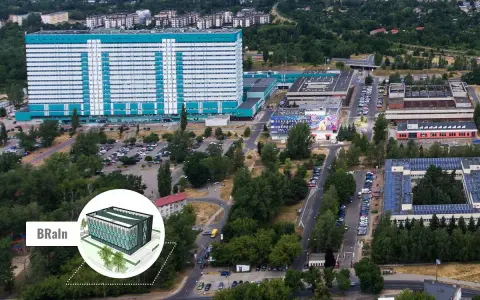 Widok z lotu ptaka na kompleks budynków, w tym duży, jasny budynek z niebieskimi akcentami, przypominający umed w Łodzi. Widać parkingi, drogi i zieleń.