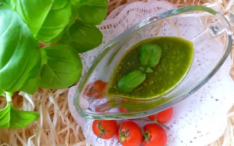Aromatyczne pesto bazyliowe w szklanej sosjerce, ozdobione listkami bazylii, obok gałązki pomidorków koktajlowych.